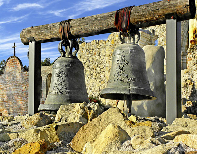 Two mission bells on a wooden frame with mission buildings in the background